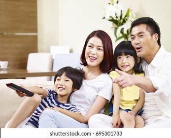 Happy Asian Family With Two Children Sitting On Sofa Watching TV Together.