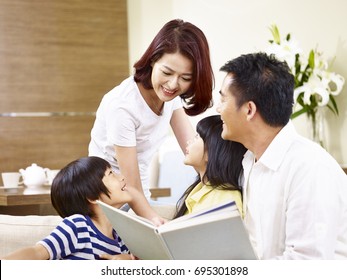 Happy Asian Family With Two Children Sitting On Sofa Reading A Book Together.