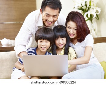 Happy Asian Family With Two Children Sitting On Couch At Home Using Laptop Computer Together.