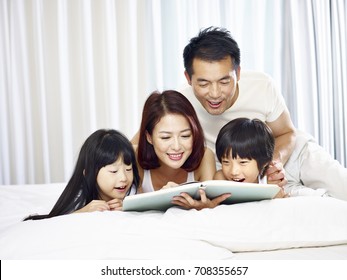 Happy Asian Family With Two Children Lying On Front In Bed Reading A Book Together.