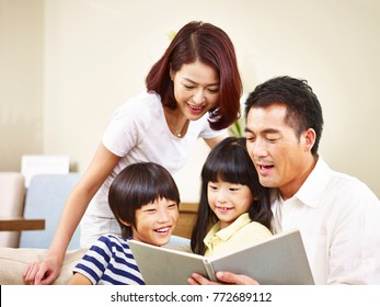 Happy Asian Family With Two Children Sitting On Sofa Reading A Book Together.