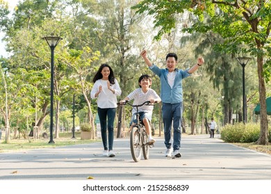 Happy Asian Father And Mother Teach Their Son To Ride A Bicycle, Cheerful Parent Raise Hands Up In The Air To Support Kid Encouragement, Family Do Activity Together At Park Concept