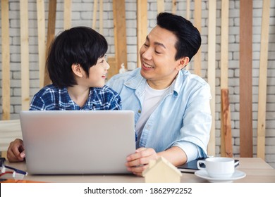 Happy Asian father and son using laptop computer and looking each other together in carpentry workshop. Concept family doing hobby together at home.