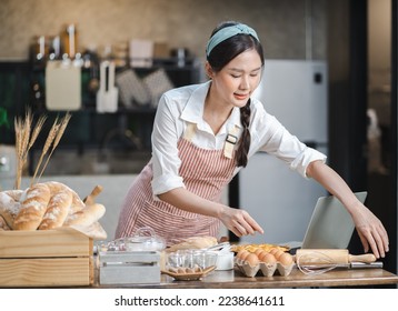 Happy Asian female baker wears apron standing at table in kitchen preparing homemade bakery ingredients. Smiling young beautiful woman baking bread and cooking at home as her small cookery business. - Powered by Shutterstock