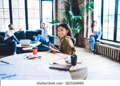 Happy Asian female in casual wear sitting at wooden table and taking notes in notebook while working with colleagues in loft office