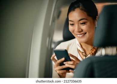 Happy Asian Female CEO Text Messaging On Mobile Phone Sitting On The Backseat Of A Car.