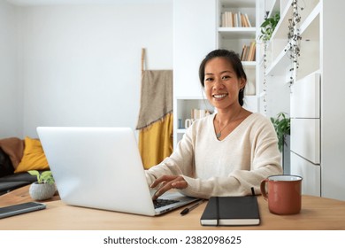 Happy Asian female entrepreneur working using laptop looking at camera. Smiling Chinese woman working with computer at home office. Technology concept.
