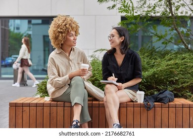 Happy Asian female journalist meets with famous person to take interview writes down notes in notebook drink takeaway coffee have pleasant conversation pose together against urban background - Powered by Shutterstock