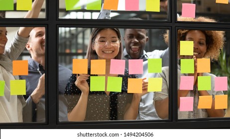Happy asian female leader and multiethnic team celebrate corporate goal achievement, completed project success, finished task result, good teamwork growth stand behind glass wall with sticky notes