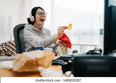happy asian female nerd holding bag of chip snack junk food with trash on desk looking monitor laughing. relax lazy teenage girl at home watch comedy movie on computer with headset sit in messy room