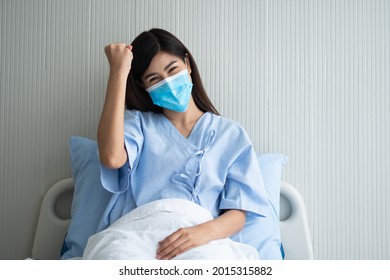 Happy Asian Female Patient Wearing A Mask, Lies On The Bed, And I Raised An Arms And Was Delighted For Show Confidence In Treatment. Concept Of Believe In Treatment And Insurance Coverage