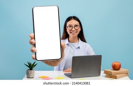 Happy asian female student demonstrating big smartphone with blank screen, showing copy space for mobile app or website ad, sitting at workplace over blue background