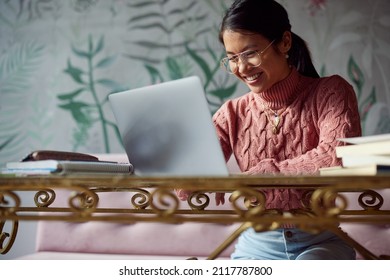 A happy Asian female student is sitting in the cafeteria and typing a report on her laptop. There are books on a table. Student exchange. - Shutterstock ID 2117787800