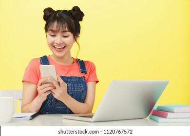 Happy Asian Female Student Using Her Mobile Phone At The Table