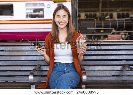 Happy Asian female traveler sits back on a chair, holding a smartphone and a cup of iced coffee in her hand, while looking for tourist attractions.