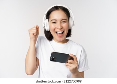 Happy Asian Girl Gamer, Playing On Mobile Phone, Watching On Smartphone, Wearing Headphones, Standing Over White Background