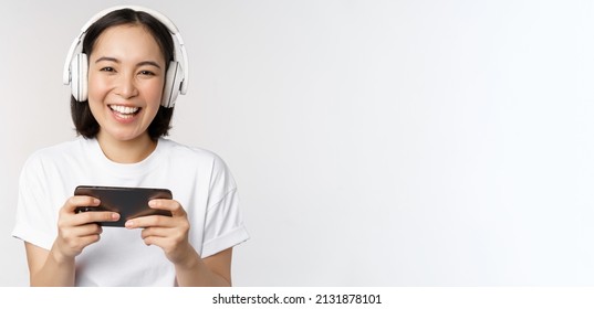 Happy Asian Girl Gamer, Playing On Mobile Phone, Watching On Smartphone, Wearing Headphones, Standing Over White Background