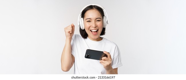 Happy Asian Girl Gamer, Playing On Mobile Phone, Watching On Smartphone, Wearing Headphones, Standing Over White Background