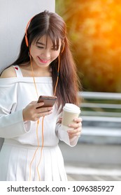 Happy asian girl with headphones listening to music from mobile phone drinking coffee. Relaxed woman holding mobile phone and coffee cup, selective focus color filter effect,copy space.