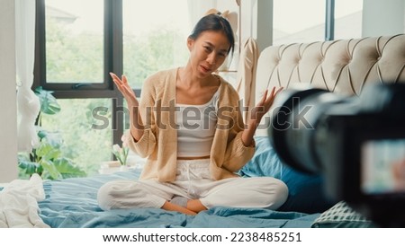 Happy Asian girl influencer with white cream pajamas recording sharing morning routine in camera sit on fluffy comfort bed talk with subscriber in bedroom morning light. Female morning vibes concept.
