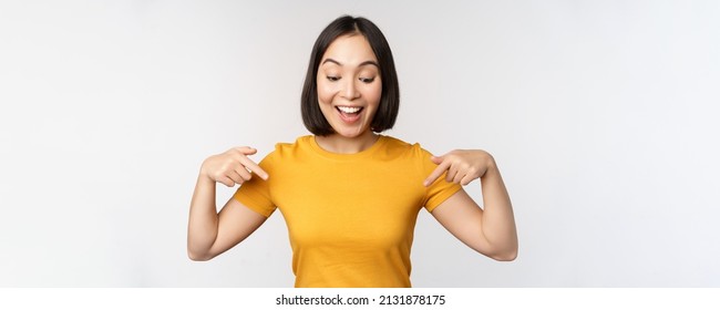 Happy asian girl pointing fingers down, looking at announcement, banner or advertisment, standing in yellow tshirt over white background