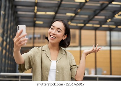 Happy asian girl shows something behind her during video call, demonstrating smth, standing on street. - Shutterstock ID 2236765819