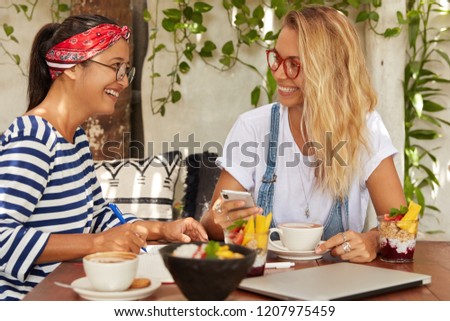 Happy Asian girl in striped sweater and eyewear makes notes in notebook, list to do, has pleasant conversation with pretty blonde young woman who reads some ideas in intenet, holds cell phone