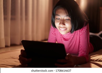  Happy Asian Girl Is Studying Online Via The Internet On Tablet While In The Bedroom In Night, Asia Child Watching And Education Distance On The Computer, Smile On The Face. Concept Learning At Home