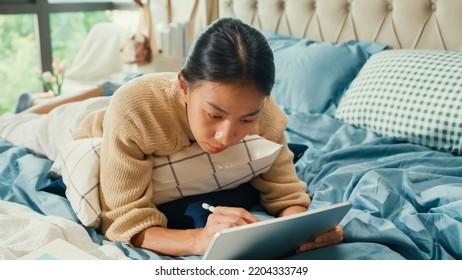 Happy Asian Girl University With White Cream Pajamas Lay Down On Fluffy Comfort Bed Use Digital Tablet Learning Online In Bedroom At Home. Revisualizing Flexible Working, Work From Home Concept.