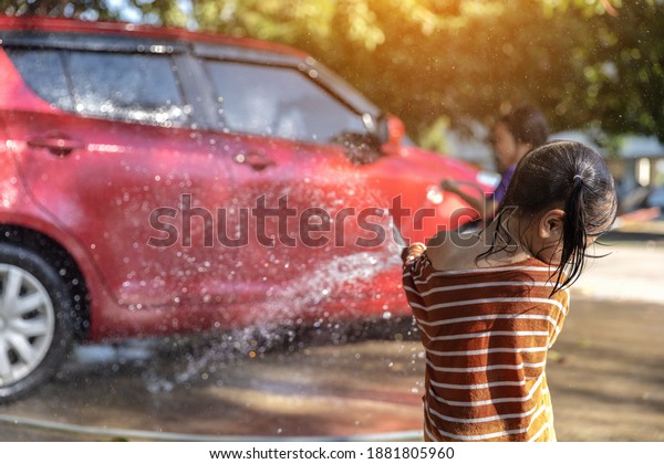 Happy Asian girl washing red car on water splashing\
and sunlight at home.