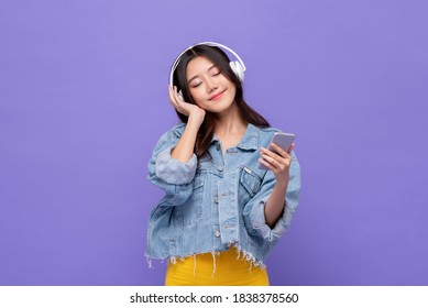 Happy Asian girl wearing headphones listening to music from mobile phone relaxing with eyes closed on isolated purple studio background - Powered by Shutterstock