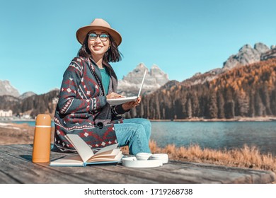 Happy Asian Girl Working At A Computer While Relaxing In A Country Nature Park. The Purest Mountain Lake In The Background