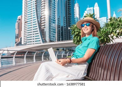 Happy asian girl works at a laptop in Dubai Marina port against the backdrop of high skyscrapers and the Bay