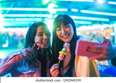 Happy Asian Girls Eating Candy Sweets And Taking Selfie At Amusement Park - Young Trendy Friends Having Fun With Technology Trend - Tech, Friendship And Influencer Concept - Focus On Right Female Face