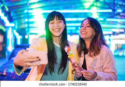 Happy Asian Girls Eating Candy Sweets And Taking Selfie At Amusement Park - Young Trendy Friends Having Fun With Technology Trend - Tech, Friendship And Influencer Concept - Focus On Left Female Face