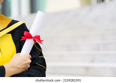 Happy Asian Graduate Student Holding The Diplomas On Hand During The University Graduation Ceremony. Master Degree Student In Gown Suit Holding A Diplomas For Photography.