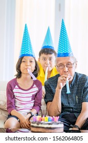 Happy Asian Grandfather And Grandchildren Blowing Horns And Wearing Party Hat At Family Birthday Party