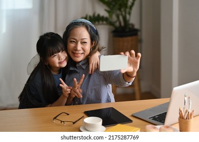 Happy Asian Grandma Making Selfie With Cute Little Granddaughter, Enjoying Carefree Leisure Weekend Activity Together In Living Room