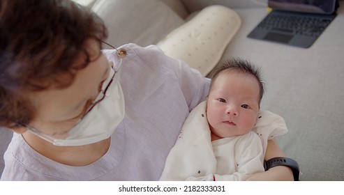 Happy Asian Grandmother With Protective Mask Talking Looking At Granddaughter Newborn Baby Infant In Grandmother Comfortable Arms At Home, Retired Grandparent Take Care Of Grandchild, Childhood	
