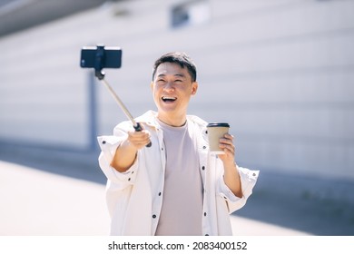 Happy asian guy makes a photo with a selfie stick on the background of a city street