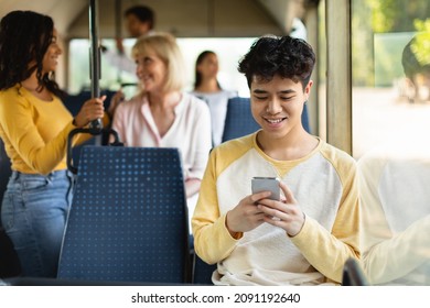 Happy Asian guy using cell playing mobile games online on smartphone connected to public wifi sitting on seat in city bus. Smiling teen passenger looking at device gadget screen, texting sms - Powered by Shutterstock