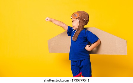 Happy Asian Handsome Funny Child Or Kid Little Boy Smile Wear Pilot Hat Play And Goggles Raise Hand Up With Toy Cardboard Airplane Wings Flying, Studio Shot Isolated Yellow Background, Startup Freedom