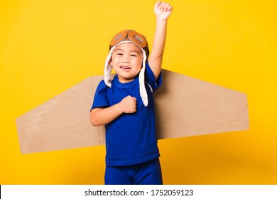 Happy Asian Handsome Funny Child Or Kid Little Boy Smile Wear Pilot Hat Play And Goggles Raise Hand Up With Toy Cardboard Airplane Wings Flying, Studio Shot Isolated Yellow Background, Startup Freedom