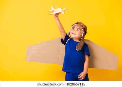 Happy Asian Handsome Funny Child Or Kid Little Boy Smile Wear Pilot Hat Play And Goggles With Toy Cardboard Airplane Wings Fly Hold Plane Toy, Studio Shot Isolated Yellow Background, Startup Freedom