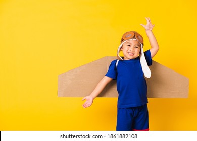 Happy Asian Handsome Funny Child Or Kid Little Boy Smile Wear Pilot Hat Play And Goggles Raise Hand Up With Toy Cardboard Airplane Wings Flying, Studio Shot Isolated Yellow Background, Startup Freedom