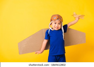 Happy Asian Handsome Funny Child Or Kid Little Boy Smile Wear Pilot Hat Play And Goggles With Toy Cardboard Airplane Wings Fly Hold Plane Toy, Studio Shot Isolated Yellow Background, Startup Freedom