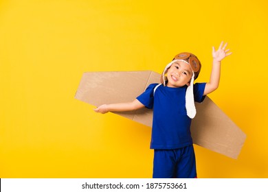 Happy Asian Handsome Funny Child Or Kid Little Boy Smile Wear Pilot Hat Play And Goggles Raise Hand Up With Toy Cardboard Airplane Wings Flying, Studio Shot Isolated Yellow Background, Startup Freedom