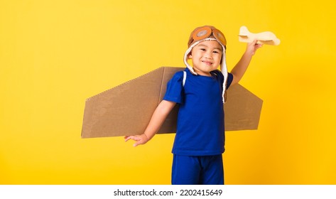 Happy Asian Handsome Funny Child Or Kid Little Boy Smile Wear Pilot Hat Play And Goggles With Toy Cardboard Airplane Wings Fly Hold Plane Toy, Studio Shot Isolated Yellow Background, Startup Freedom