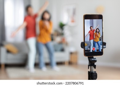 Happy Asian Husband And Wife Recording Video For Social Media Or Personal Family Vlog, Holding Hands And Dancing In Front Of Camera, Selective Focus On Smartphone Set On Tripod, Home Interior