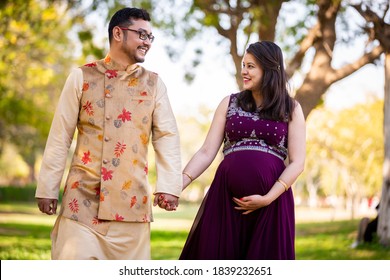 Happy Asian Indian Pregnant Woman With Her Husband In Walking Outdoor In A Park Or Garden, Smiling Cheerful Young Parents Looking At Camera Expecting Baby.